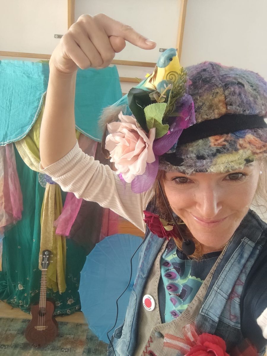 Araceli Lozano caracterizada de cuentacuentos, señalando el pájaro de su sombrero de flores, con telas de colores y un ukelele al fondo