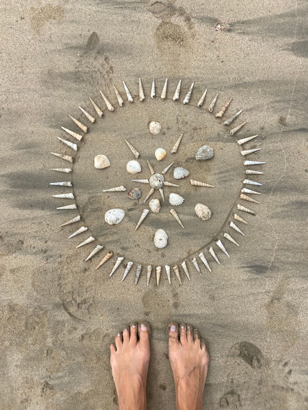 Mandala de conchas sobre la arena, con unos pies descalzos ante él