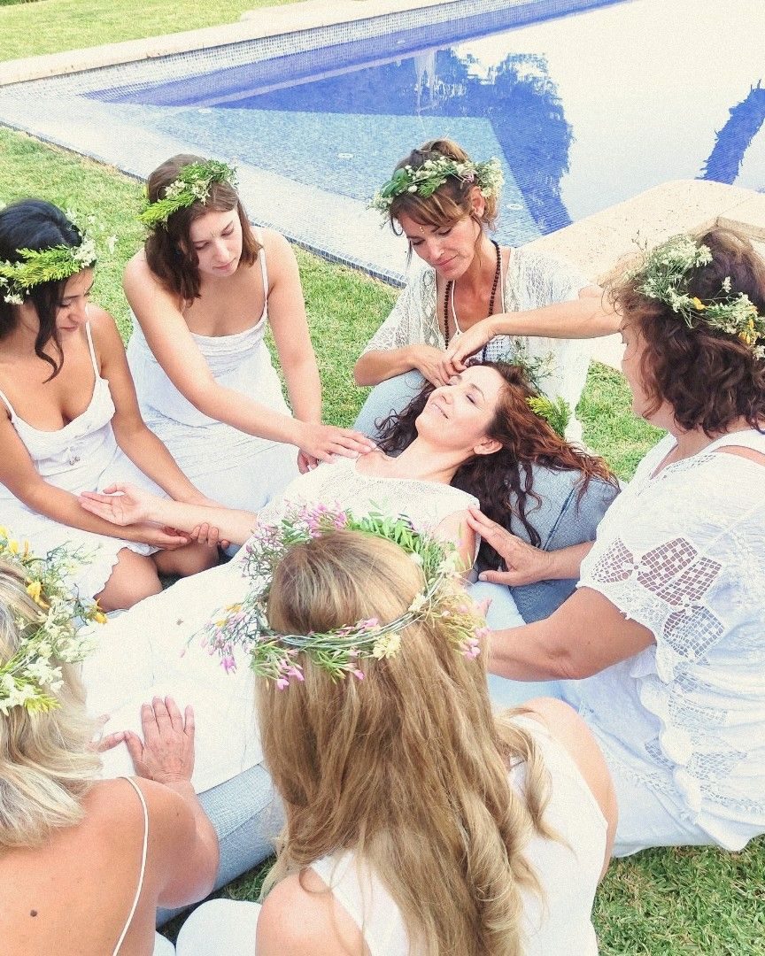 Mujeres con coronas de flores posan sus manos sobre una compañera tumbada, junto a la piscina